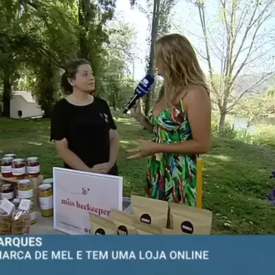 Produtos de mel em frascos num expositor ao ar livre com flores e duas mulheres conversando