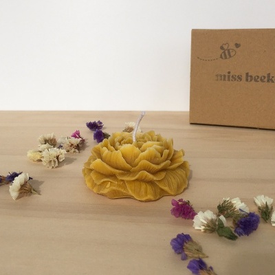 Vela amarela em forma de flor com flores secas ao redor e caixa de papelão ao fundo