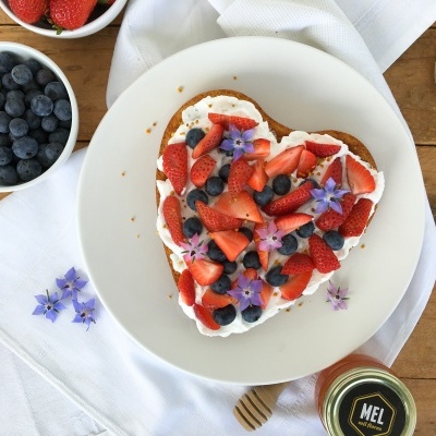 Receita Bolo Dia da Mãe Torrada em forma de coração com frutas e flores comestíveis num prato branco sobre mesa de madeira