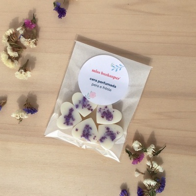 Cinco velas em forma de coração com flores roxas em embalagem transparente sobre mesa de madeira com flores secas ao redor