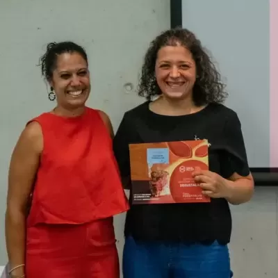 Duas mulheres sorridentes junto a quadro branco, uma com vestido vermelho, outra com blusa preta e calças azuis segurando caixa vermelha.