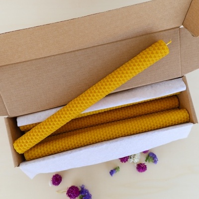 Velas amarelas de cera de abelha em caixa de papelão com forro branco e flores secas ao redor.