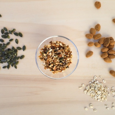 Tigela de granola com sementes de abóbora, amêndoas e aveia ao redor sobre madeira clara