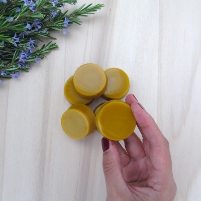 Bola de cera amarelo-dourada com uma mão segurando e erva com flores roxas sobre fundo de madeira clara