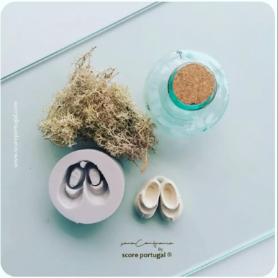 Molde rosa com sapatos minúsculos, sapatos brancos, musgo seco e frasco verde com tampa de cortiça sobre superfície branca