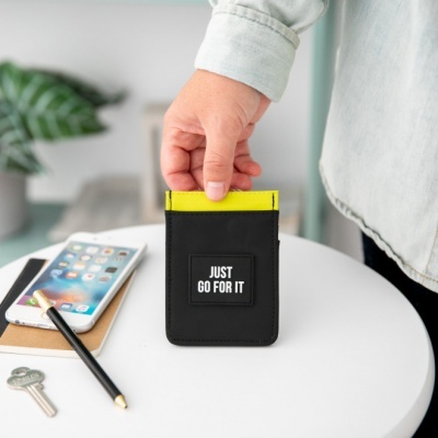 Carteira preta com texto JUST GO FOR IT, mão segurando, mesa branca com chave, lápis, bloco de notas e smartphone