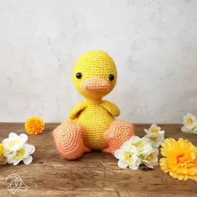 Boneco de pato em crochet amarelo e laranja com flores ao redor