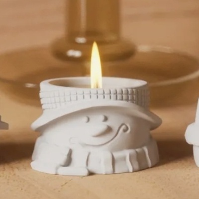 Três velas decorativas brancas acesas com suportes em forma de personagens sorridentes.