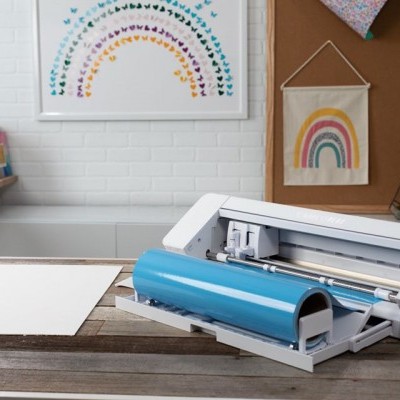 Impressora de vinil branca com rolo azul numa mesa de madeira, com decoração de arco-íris na parede