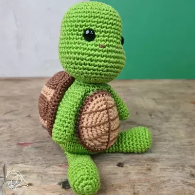 Boneco de tartaruga em croché verde, castanho e bege, sentado numa mesa de madeira