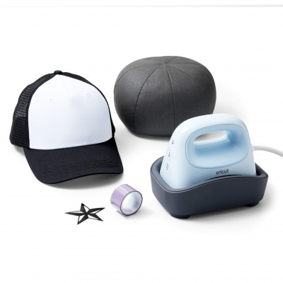 Boné preto e branco, estojo preto redondo, fita lilás, enfeite estrela preto e ferro de engomar azul claro com base preta Cricut