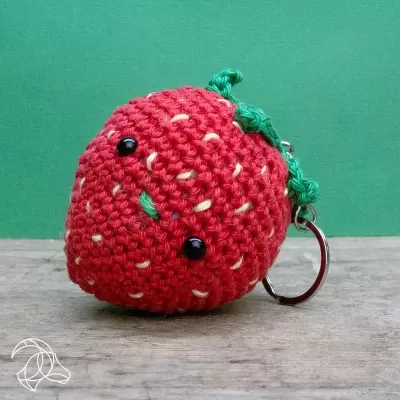 Porta-chaves em crochê vermelho e verde em forma de morango com olhos de botão pretos sobre superfície de madeira