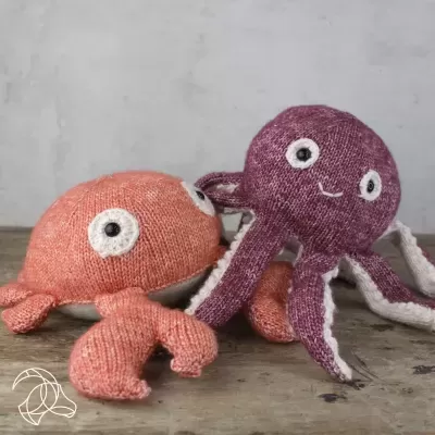 Dois brinquedos de pelúcia tricotados, um caranguejo laranja e um polvo roxo, sobre madeira