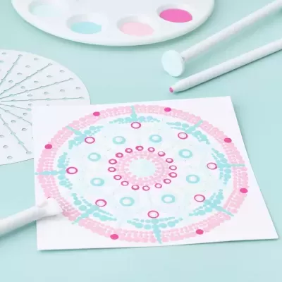 Conjunto para pintura de mandalas com pincel, paleta de tintas e base com padrão circular azul e rosa.