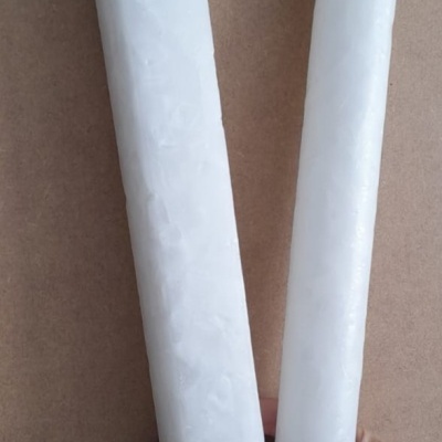 Duas velas brancas, uma quadrada e uma cilíndrica, seguradas por uma mão com unhas pintadas, fundo castanho