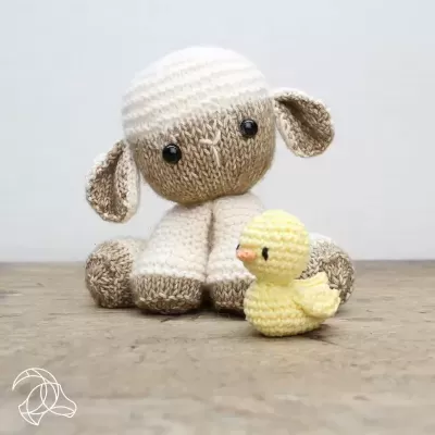Peluches tricotados de cordeiro creme e castanho claro e patinho amarelo com olhos pretos grandes sobre chão de madeira
