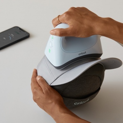 Dispositivo eletrónico Cricut EasyPress Mini a aplicar calor num boné cinza claro