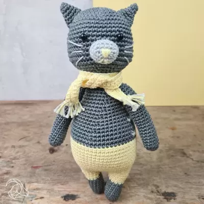 Boneco croché de gato cinzento com cachecol amarelo em superfície de madeira