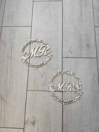 Dois enfeites de madeira com letras MR e MRS em círculos com padrão de ramos sobre piso de madeira cinzenta