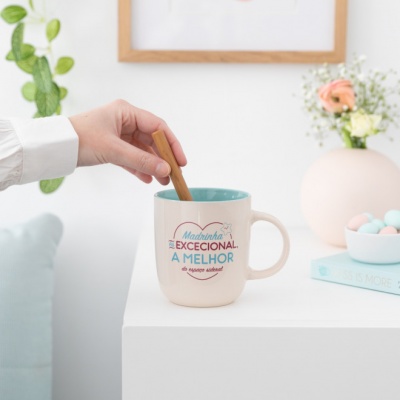 Caneca com texto colorido sobre mesa branca, com mão a mexer e decoração floral