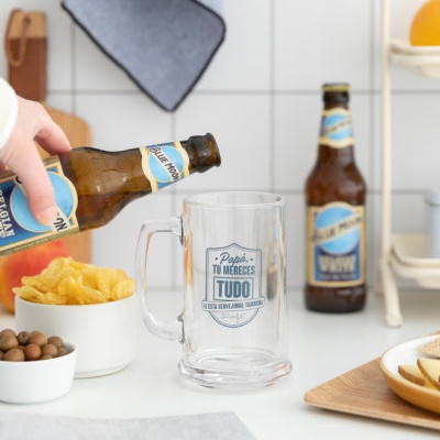 Pessoa a verter cerveja numa caneca transparente com texto azul, garrafa com rótulo azul e branco e petiscos em fundo de cozinha