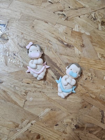 Dois bonecos de bebés com chupetas, um azul e outro rosa, sobre superfície de madeira clara