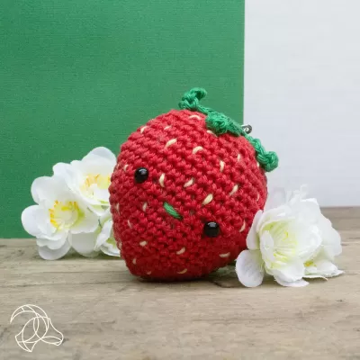 Amigurumi de morango vermelho em crochê com olhos e boca, ao lado de flores brancas sobre madeira.