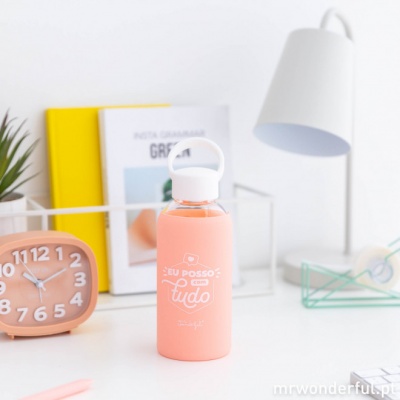 Garrafa de água cor de salmão com texto branco ao centro, sobre mesa branca com objetos em fundo
