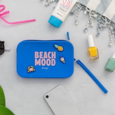 Bolsa azul com texto BEACH MOOD e pins decorativos rodeada por cosméticos, telemóvel, e acessórios de praia
