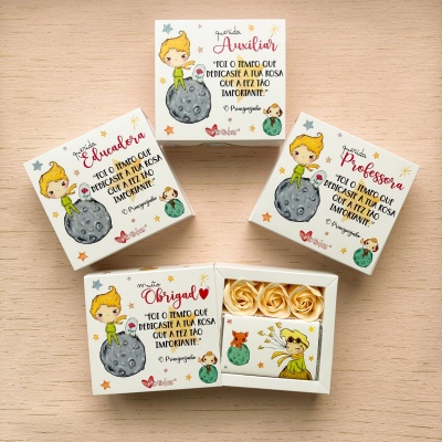 Caixas de papel decorativas com ilustrações e texto do Pequeno Príncipe, flores artificiais e sabonete em fundo de madeira clara.