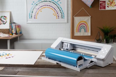 Impressora de vinil branca com rolo azul numa mesa de madeira, com decoração de arco-íris na parede