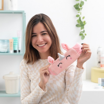 Máscara de dormir rosa com desenho de unicórnio, segurada por mulher sorridente em ambiente claro