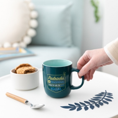 Caneca azul petróleo com texto amarelo, em mesa branca com bolachas e colher