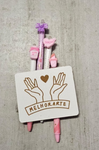 Três esferográficas cor-de-rosa com decoração em cima e cartão branco com desenho de mãos e a palavra MELHORARTE