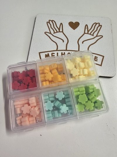 Pequenos objetos em forma de flor em caixa plástica sobre suporte branco com desenho de mãos e coração e texto 'MELHOR AMIGO'.