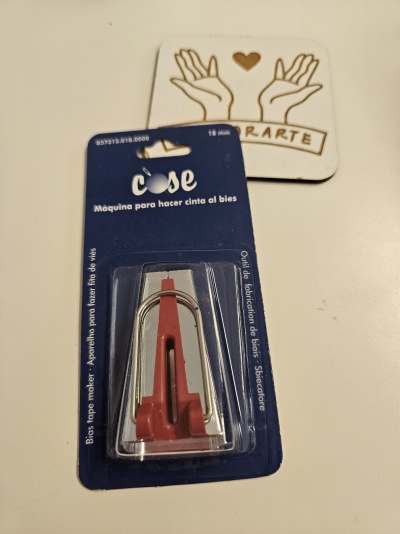 Ferramenta vermelha e metalizada para fita ao viés em embalagem azul e transparente, junto a suporte branco com desenho de mãos e coração