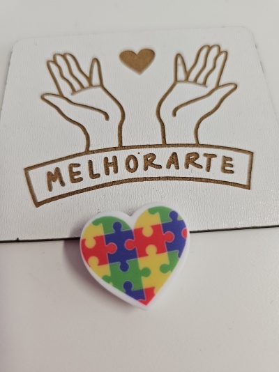Broche coração com padrão de peças de puzzle coloridas sobre cartão branco com desenho de mãos e texto MELHORARTE