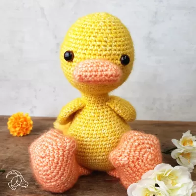 Boneco de pato amarelo em crochê com bico e pés cor de pêssego sentado com flores ao redor