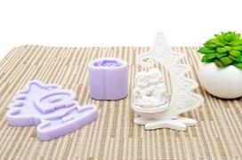 Três moldes de silicone lilás e uma escultura branca numa superfície têxtil bege às riscas com uma planta verde