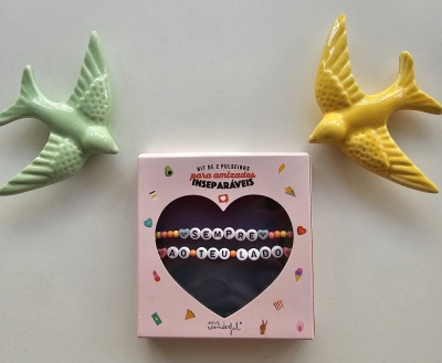 Conjunto de pulseiras coloridas em caixa rosa com janela em forma de coração, entre figuras de pássaros cerâmicos verde e amarelo