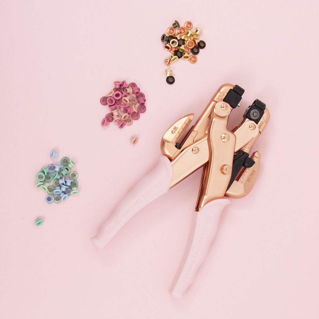 Crop-A-Dile | Ilhós | Rosa Gold Alicate cortante cobre com pegas brancas e ilhós coloridos sobre fundo rosa claro