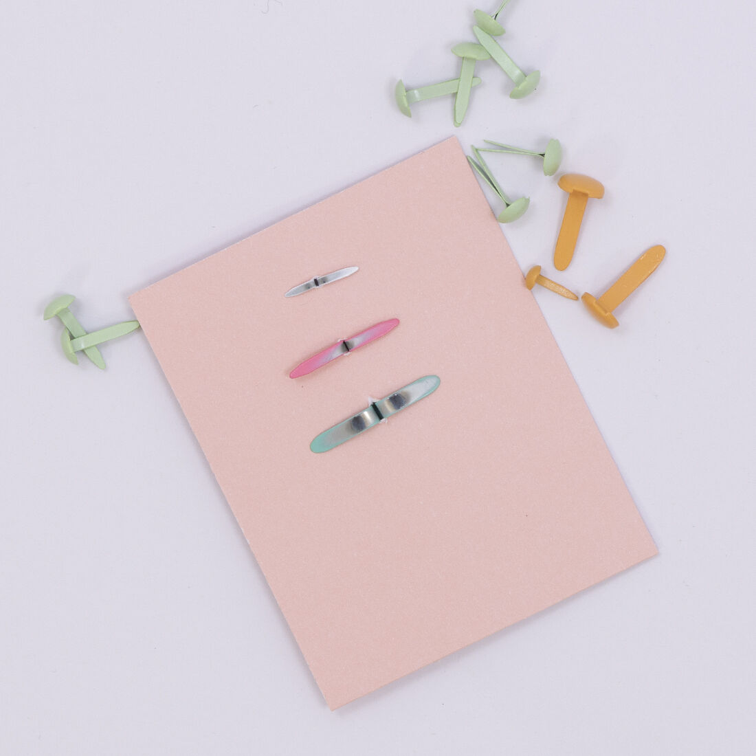 Bloco de papel rosa com três clipes metálicos coloridos e clipes de plástico verde e amarelo ao redor