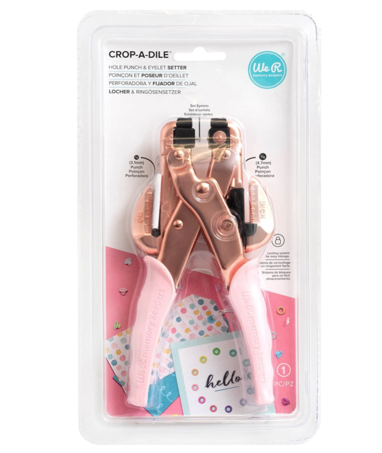 Crop-A-Dile | Ilhós | Rosa Gold Perfurador e fixador de ilhós rosa em embalagem plástica transparente com textos e cartões coloridos ao fundo.