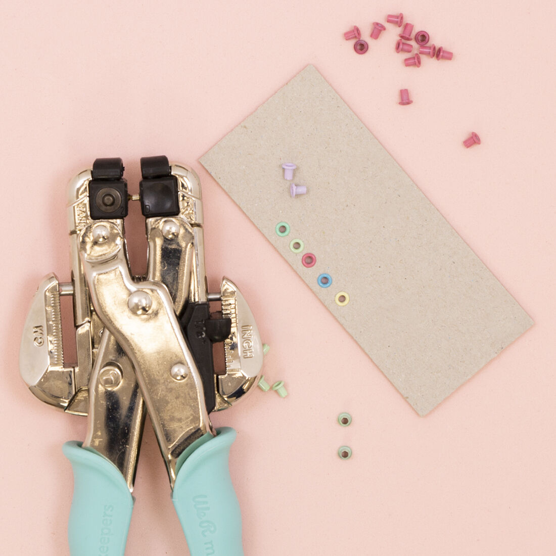 Buril metálico com punhos azuis e ilhós coloridos sobre fundo rosa