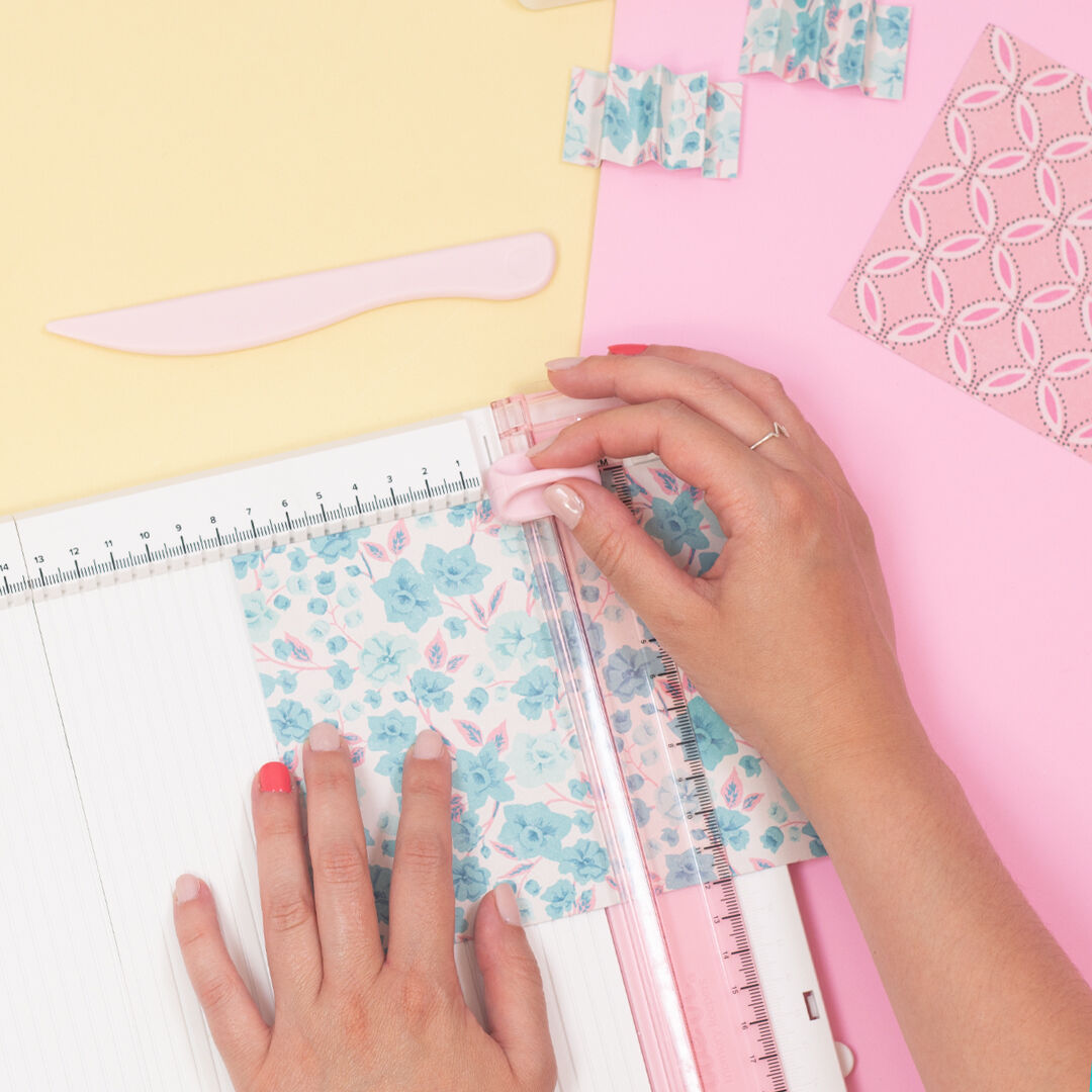 Mãos cortando papel decorativo floral com ferramenta de corte rosa