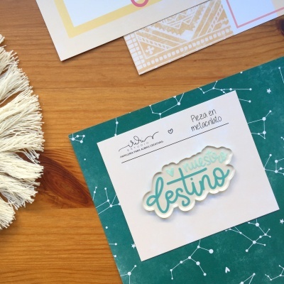 Pin metacrílico com texto turquesa e branco sobre cartão branco e superfície de madeira clara com decoração têxtil e papéis ao fundo