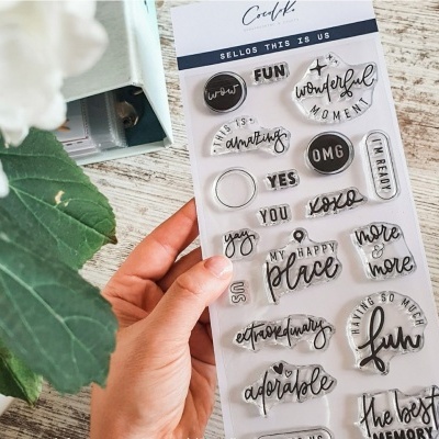 Conjunto de carimbos transparentes com palavras e frases em inglês segurado por uma mão sobre mesa de madeira clara