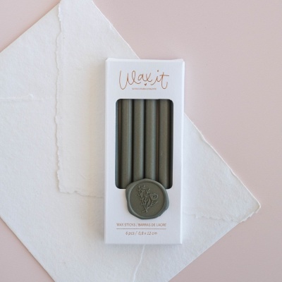 Caixa branca com seis velas cinza e selo de flor, sobre papel branco e fundo rosa claro.