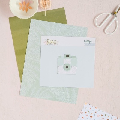 Materiais de artesanato sobre superfície clara: tesoura, fios de bordar, papéis decorativos, cartão com ilustração de câmara e tigela com recortes.