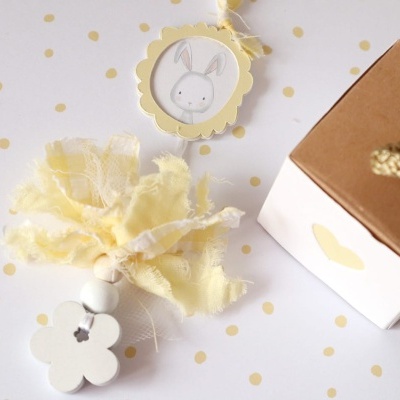Marcador decorativo com flor branca, contas e tecido amarelo claro junto a caixa de papelão